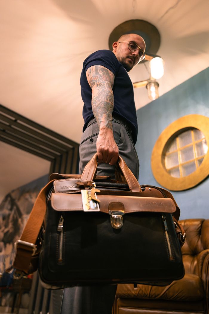 A fashionable man with tattoos holds a leather bag indoors, showcasing modern style and confidence.