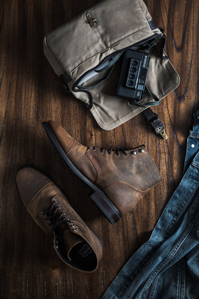 Flat lay of vintage items including leather boots, a camera, denim, and bag on wooden surface.